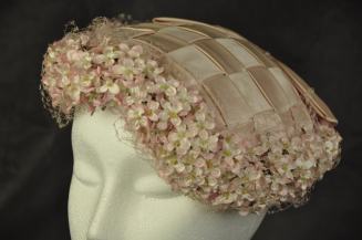 016/017 Women’s hat. Pale pink woven satin toque edged in small matching pink flowers under matching pink fascinator lace. Large pink, flat, satin bow at back of hat. Lined with satin lining material and labeled “The TLB Hat”.